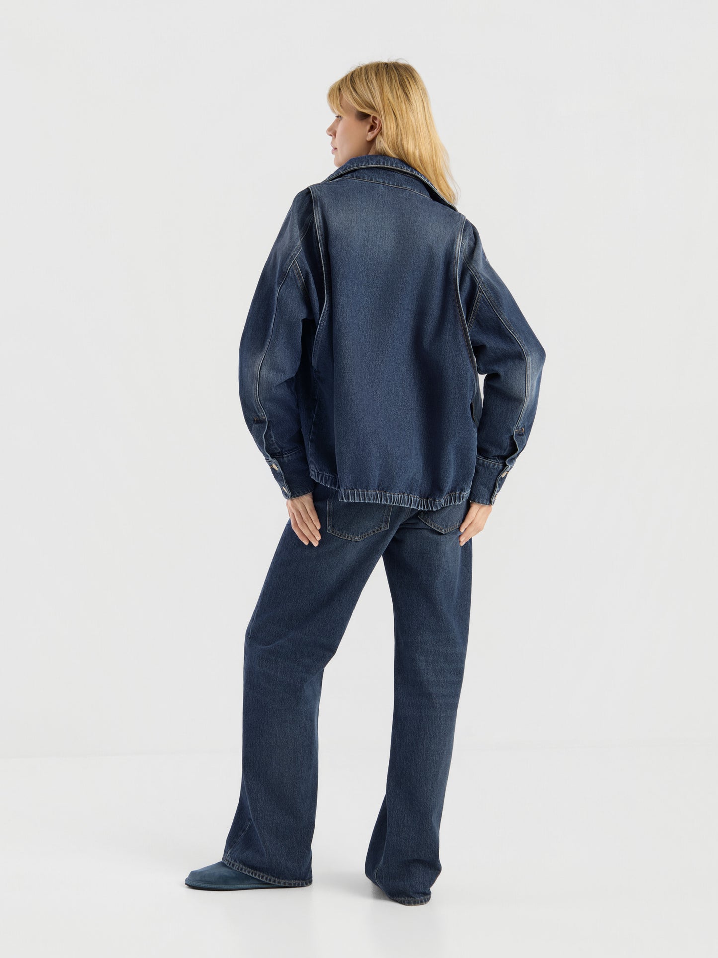 Eine blonde Frau, die mit dem Rücken zur Kamera steht, präsentiert die Oversized Denim Bomber Jacke in Dunkelblau zusammen mit einer passenden Jeans vor einem schlichten weißen Hintergrund.