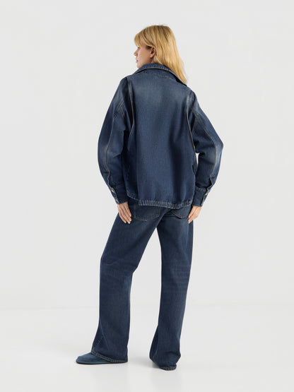 Eine blonde Frau, die mit dem Rücken zur Kamera steht, präsentiert die Oversized Denim Bomber Jacke in Dunkelblau zusammen mit einer passenden Jeans vor einem schlichten weißen Hintergrund.