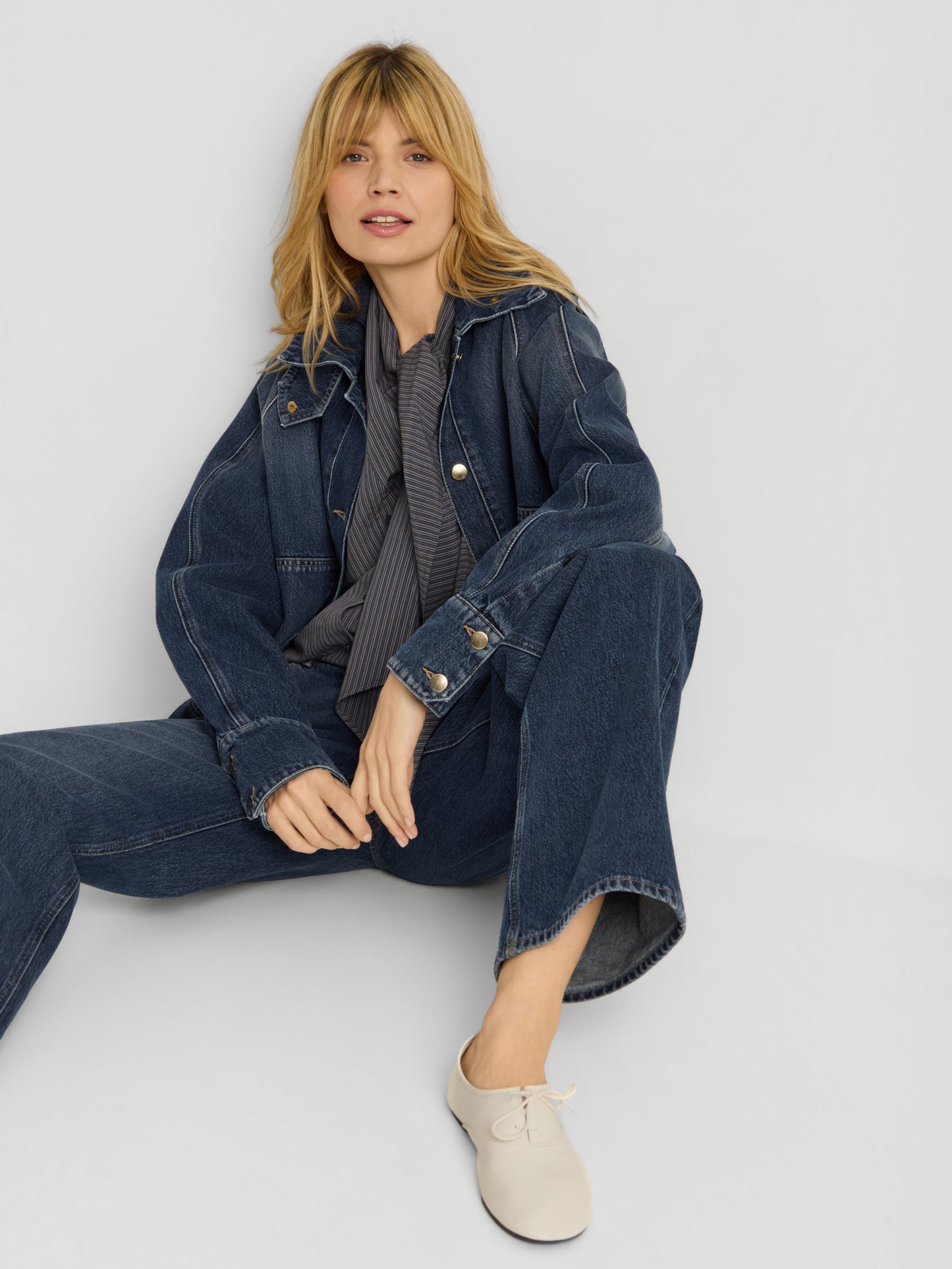 Eine blonde Frau sitzt vor einem hellen Hintergrund auf dem Boden und trägt die Oversized Denim Bomber Dark Blue, Jeans, ein dunkel gestreiftes Hemd und beige Schnürschuhe. Mit entspanntem Gesichtsausdruck blickt sie nach vorne.