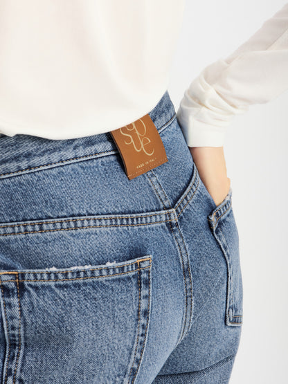 Eine Person trägt eine hoch taillierte SIENNA Jeans Light Blue – Straight Leg Damenjeans in hellblauer Waschung, kombiniert mit einem weißen Langarm-Oberteil, und hat eine Hand in die Gesäßtasche gesteckt. Ein braunes Etikett am Bund mit der Aufschrift „SUTJE“ unterstreicht die Premium-Denim-Qualität dieser Jeans.