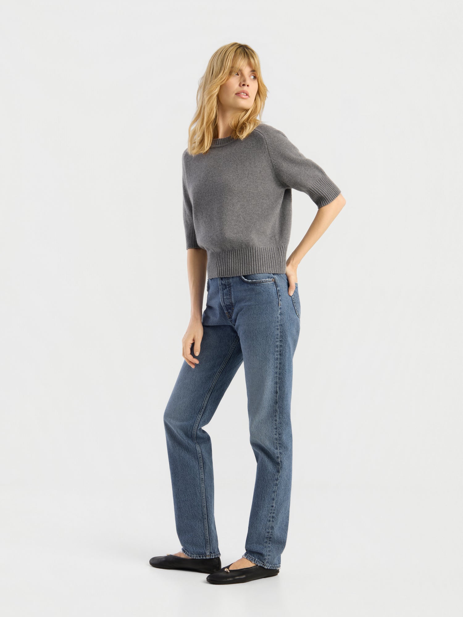 Eine Frau mit blonden Haaren steht vor einem schlichten weißen Hintergrund, eine Hand in der Tasche, und trägt einen grauen Kurzarmpullover, schwarze Ballerinas und die hochwertige SIENNA Jeans Light Blue – Straight Leg Damenjeans.
