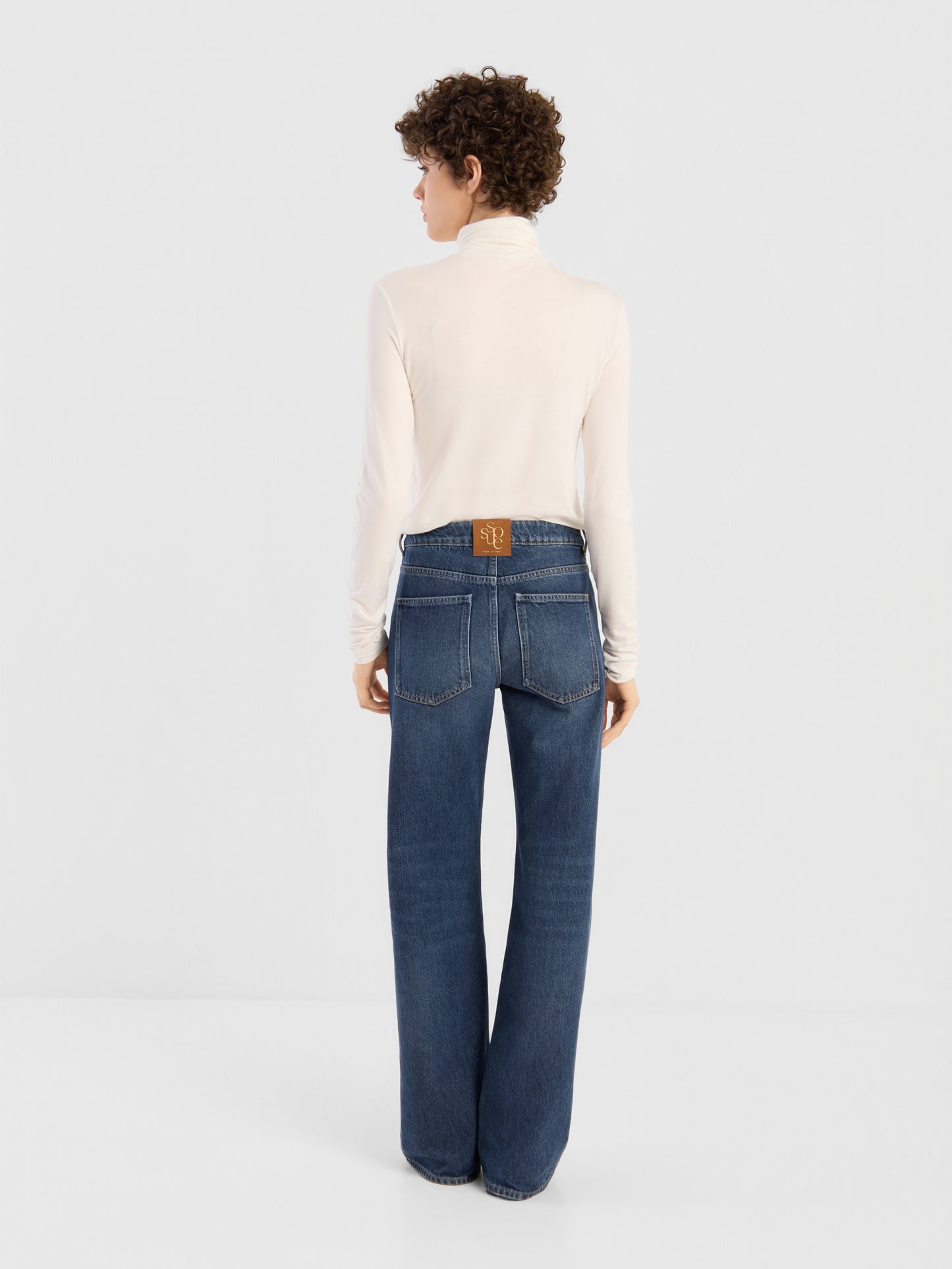 Eine Person steht mit dem Rücken zur Kamera, bekleidet mit einem weißen Rollkragenpullover und der NOLA Jeans „Dark Blue – Wide Leg“ für Damen, die sich durch sichtbare Gesäßtaschen und einen braunen Aufnäher oberhalb des Bunds auszeichnet. Diese aus Premium-Denim gefertigte Jeans verleiht der lockeren Silhouette einen modernen Touch. Der Hintergrund ist einfarbig und hell.
