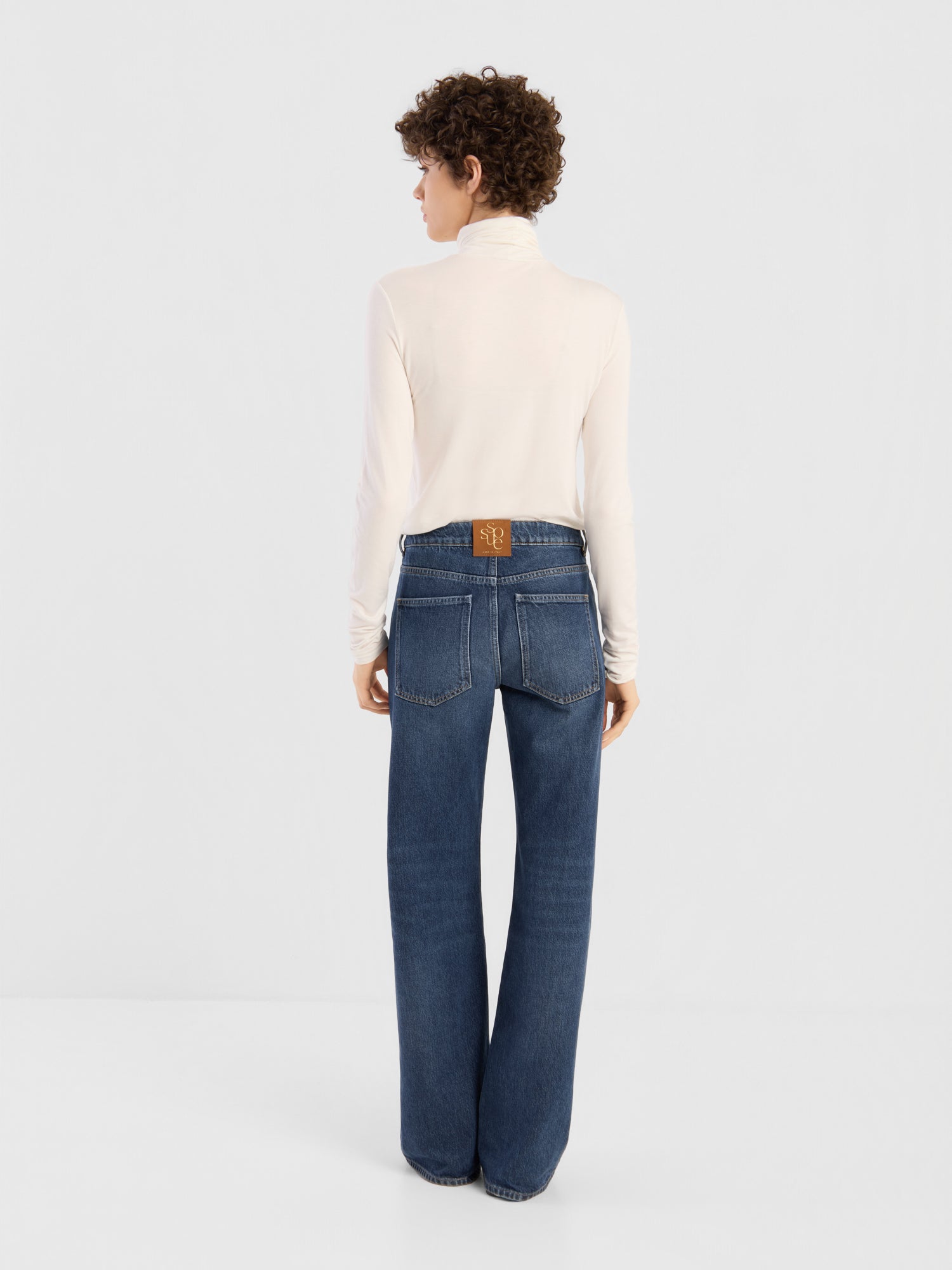 Eine Person steht mit dem Rücken zur Kamera, bekleidet mit einem weißen Rollkragenpullover und der NOLA Jeans „Dark Blue – Wide Leg“ für Damen, die sich durch sichtbare Gesäßtaschen und einen braunen Aufnäher oberhalb des Bunds auszeichnet. Diese aus Premium-Denim gefertigte Jeans verleiht der lockeren Silhouette einen modernen Touch. Der Hintergrund ist einfarbig und hell.