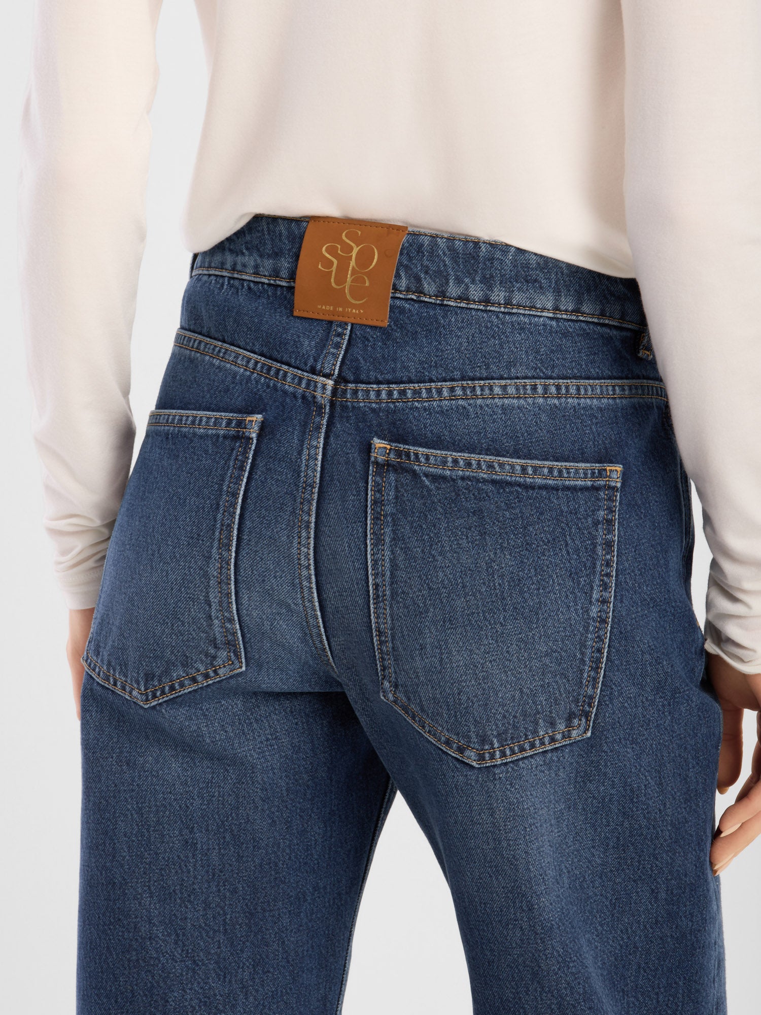 Eine Person ist von hinten zu sehen. Sie trägt ein helles Langarmhemd und dazu die NOLA Jeans Dark Blue – Wide Leg Damenjeans, die aus hochwertigem Denim gefertigt ist und somit Komfort und Langlebigkeit bietet. Dieses Modell verfügt über zwei Gesäßtaschen und ein braunes Lederaufnäher mit geprägter Schrift am Bund.
