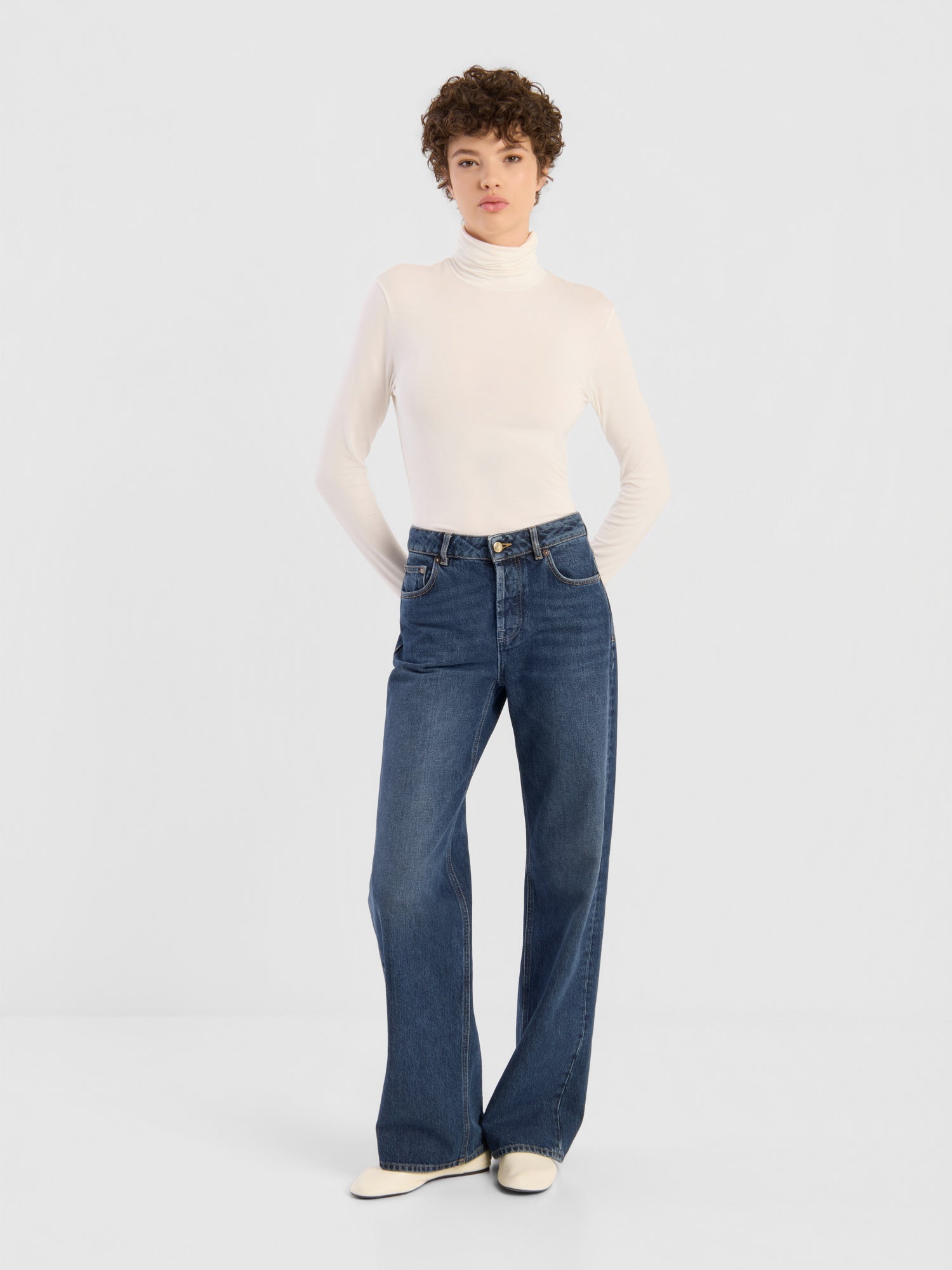 Eine Person mit kurzen Locken trägt einen weißen Rollkragenpullover, eine dunkelblaue NOLA-Jeans mit weitem Bein (Damenjeans) und weiße Schuhe und steht mit hinter dem Rücken verschränkten Händen vor einem schlichten, hellen Hintergrund.