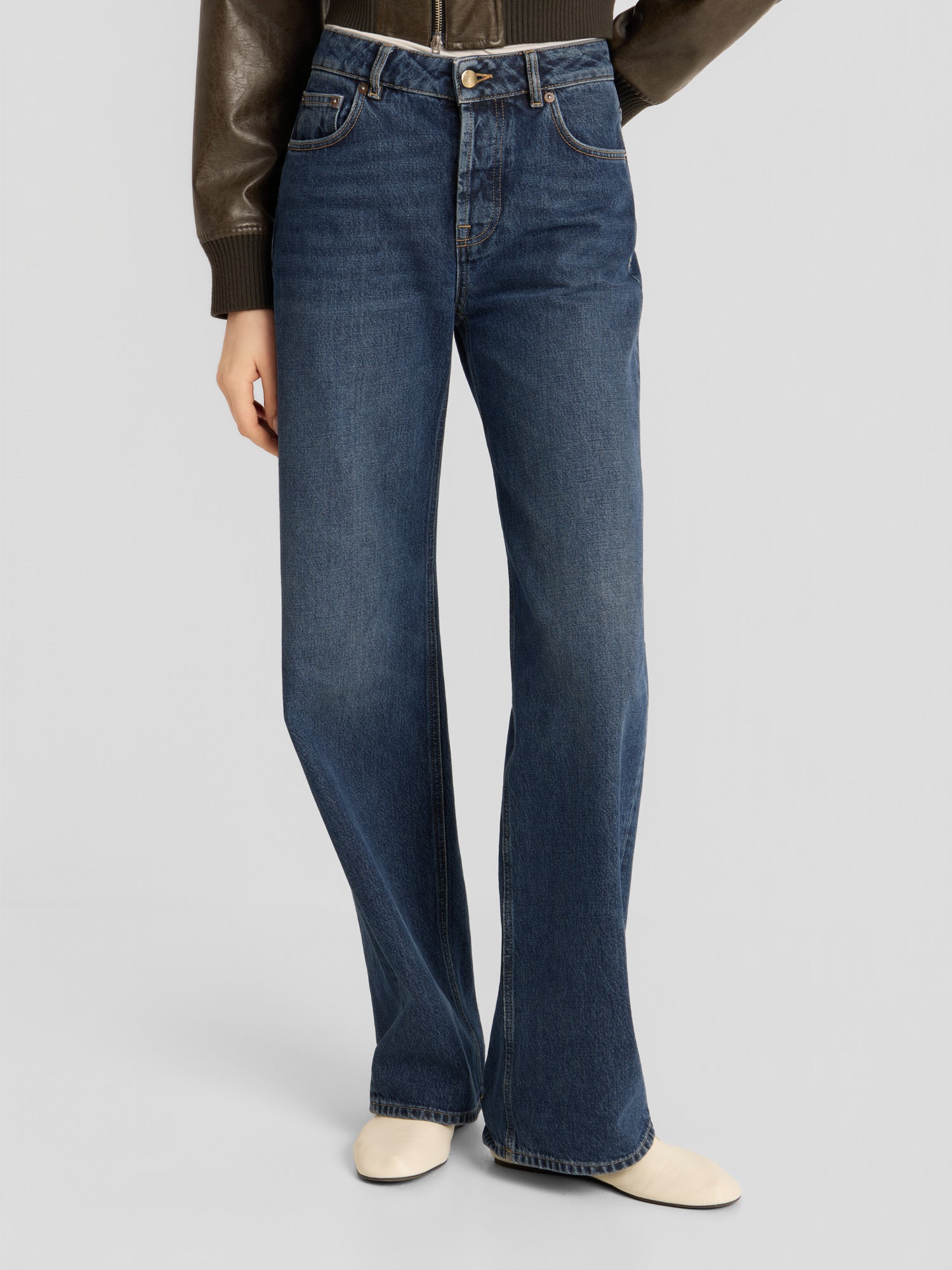 Eine Person trägt die NOLA Jeans „Dark Blue – Wide Leg“ (Damenjeans), kombiniert mit einer braunen Jacke und cremefarbenen Schuhen, und steht vor einem schlichten, hellen Hintergrund.