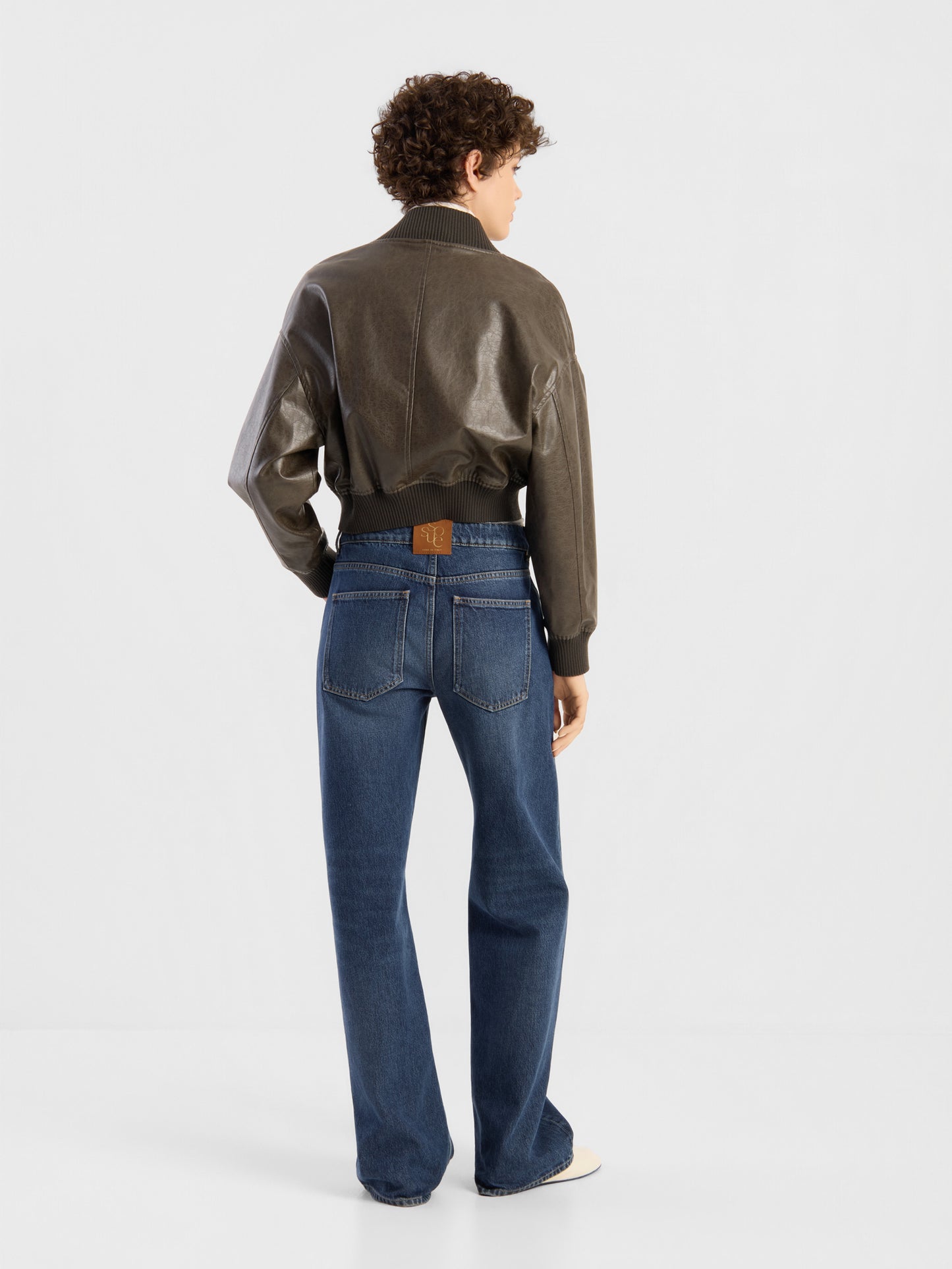 Eine Frau mit kurzen Locken und einer braunen Lederjacke trägt die NOLA Jeans „Dark Blue – Wide Leg“ (Damenjeans) und steht mit dem Rücken zur Kamera vor einem schlichten weißen Hintergrund.