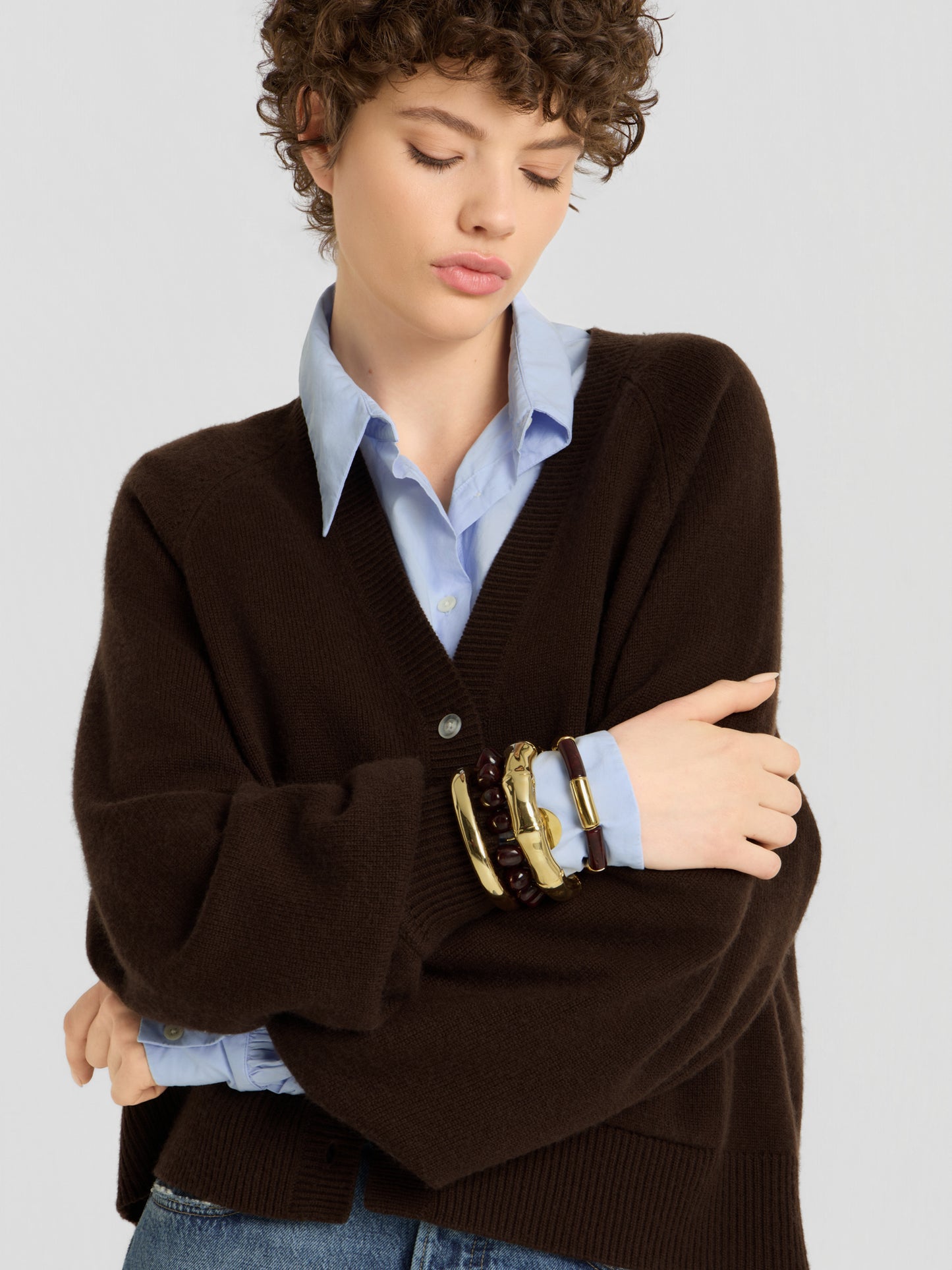 Eine Person mit kurzen Locken trägt den „Bella Cardigan Braun“ über einem blauen Hemd mit Kragen, dazu klobige goldene und dunkle Armbänder. Sie steht mit verschränkten Armen da und blickt nachdenklich nach unten.