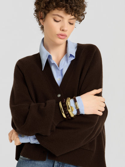Eine Person mit kurzen Locken trägt den „Bella Cardigan Braun“ über einem blauen Hemd mit Kragen, dazu klobige goldene und dunkle Armbänder. Sie steht mit verschränkten Armen da und blickt nachdenklich nach unten.