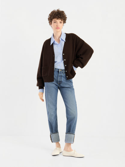 Eine Person mit kurzen Locken trägt den „Bella Cardigan Braun“ über einem hellblauen Hemd, einer blauem Jeans mit umgeschlagenen Hosenbeinen und weißen Schuhen und steht vor einem schlichten weißen Hintergrund.