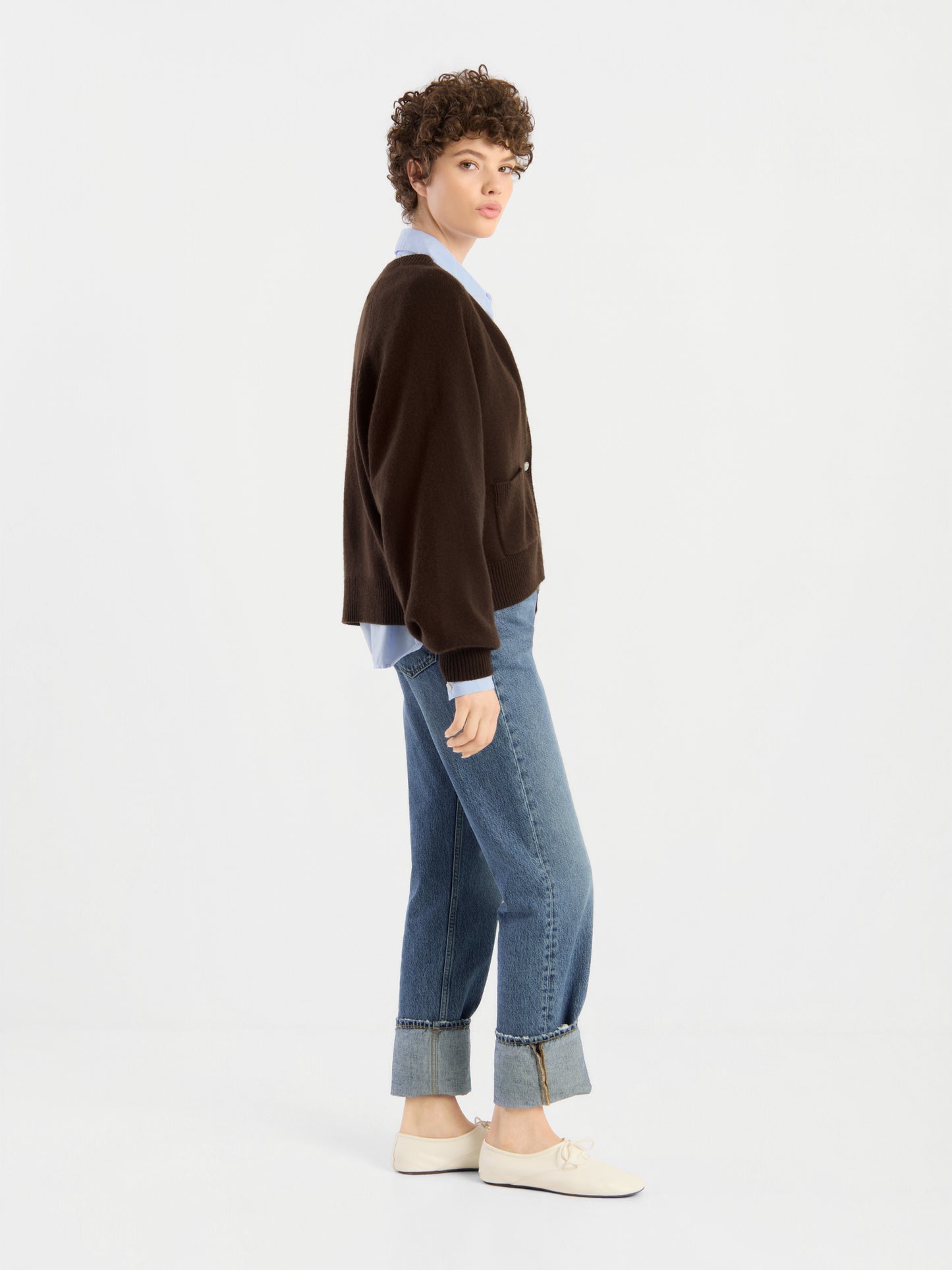Vor einem schlichten weißen Hintergrund steht eine Person mit kurzen Locken seitlich im Bella Cardigan Braun, einem hellblauen Hemd, blauen Jeans mit umgeschlagenen Beinen und weißen Sneakers.