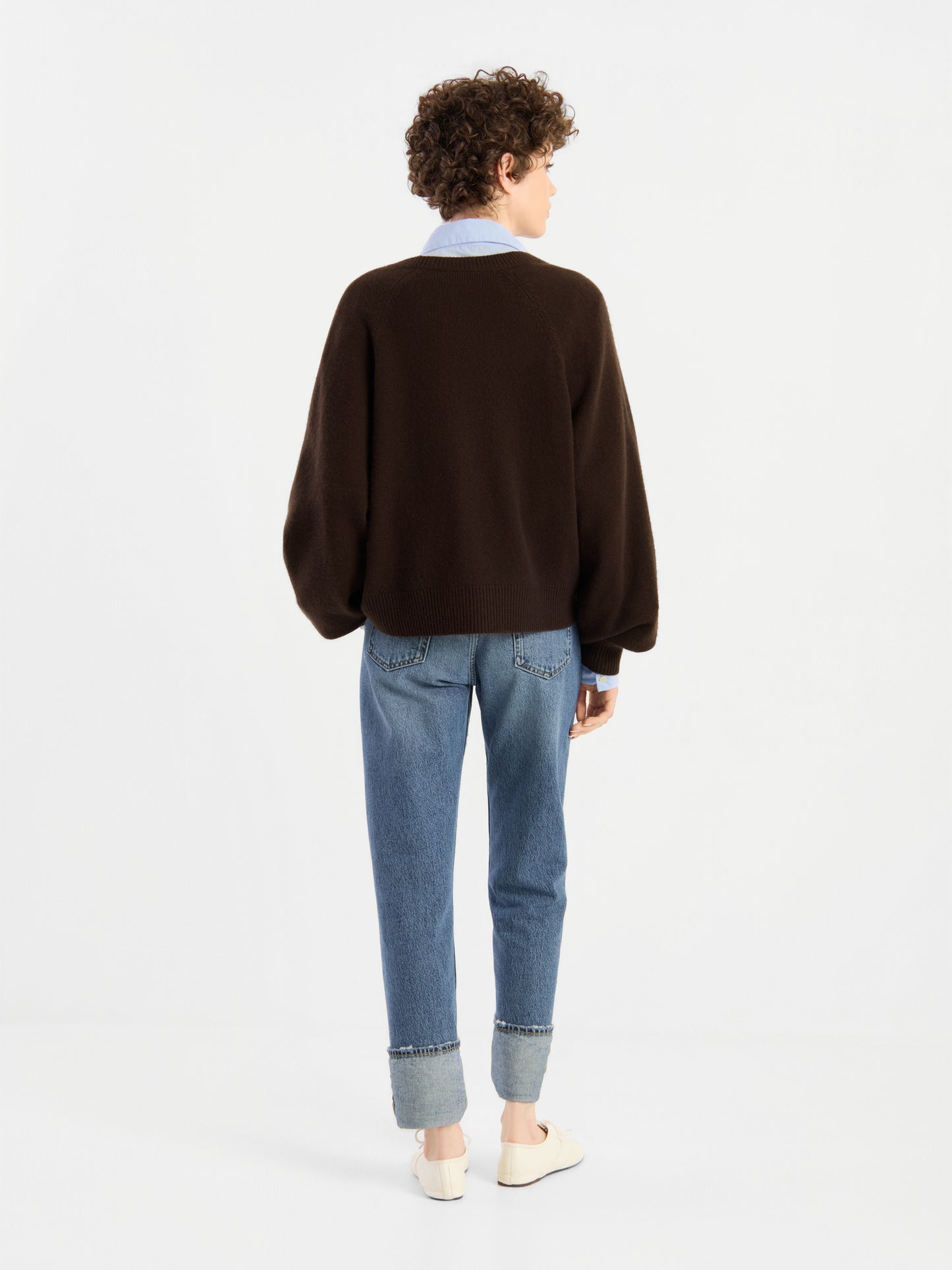 Eine Person mit kurzen Locken steht mit dem Rücken zur Kamera und trägt den „Bella Cardigan Braun“ über einem Hemd mit Kragen, dazu hellblaue Jeans mit Umschlag und weiße Schuhe vor einem schlichten weißen Hintergrund.