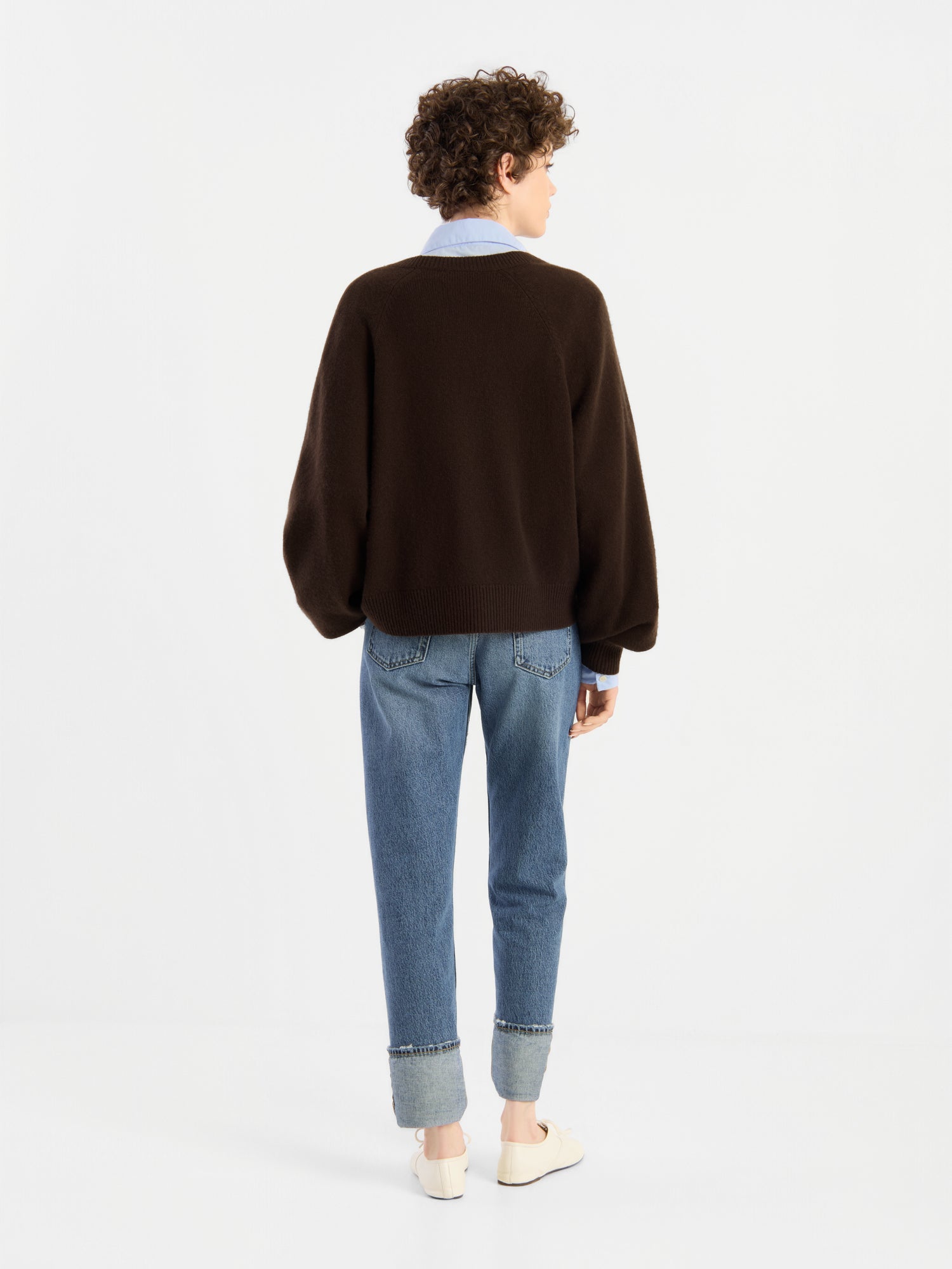 Eine Person mit kurzen Locken steht mit dem Rücken zur Kamera und trägt den „Bella Cardigan Braun“ über einem Hemd mit Kragen, dazu hellblaue Jeans mit Umschlag und weiße Schuhe vor einem schlichten weißen Hintergrund.