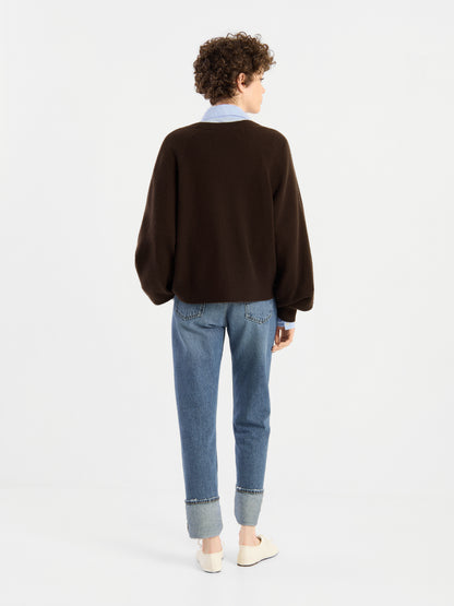 Eine Person mit kurzen Locken steht mit dem Rücken zur Kamera und trägt den „Bella Cardigan Braun“ über einem Hemd mit Kragen, dazu hellblaue Jeans mit Umschlag und weiße Schuhe vor einem schlichten weißen Hintergrund.