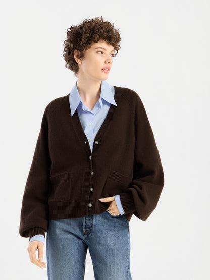 Eine Person mit kurzen Locken trägt den „Bella Cardigan Braun“ über einem hellblauen Hemd mit Kragen und einer blauen Jeans und steht vor einem schlichten weißen Hintergrund.