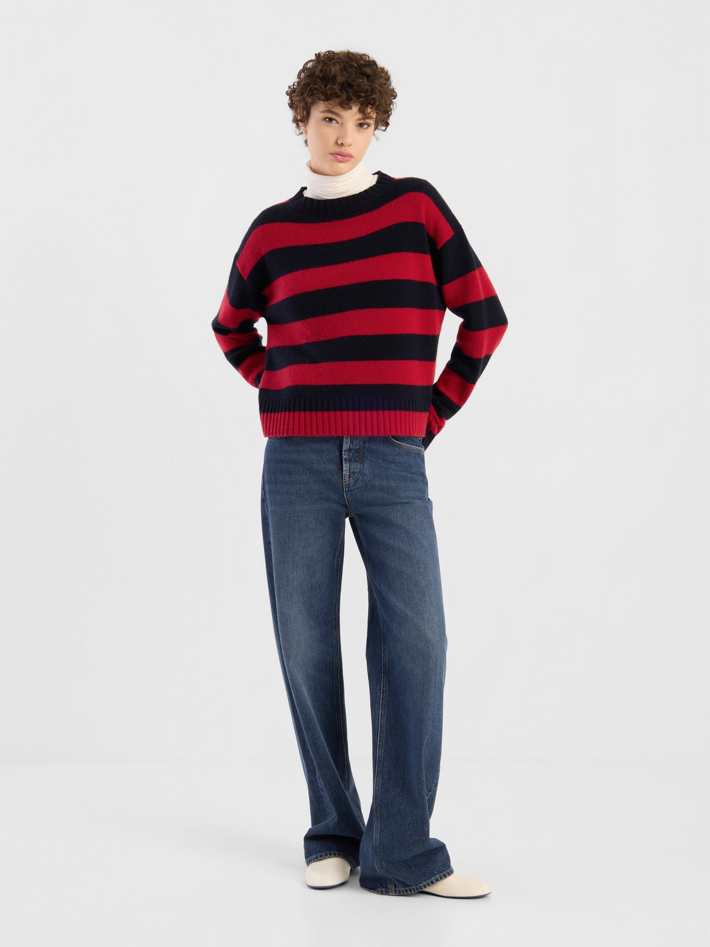 Vor einem schlichten weißen Hintergrund steht eine Person mit den Händen in den Taschen und trägt den Pullover „Red Blue Stripes“, eine blaue Jeans mit weitem Bein und weiße Schuhe.