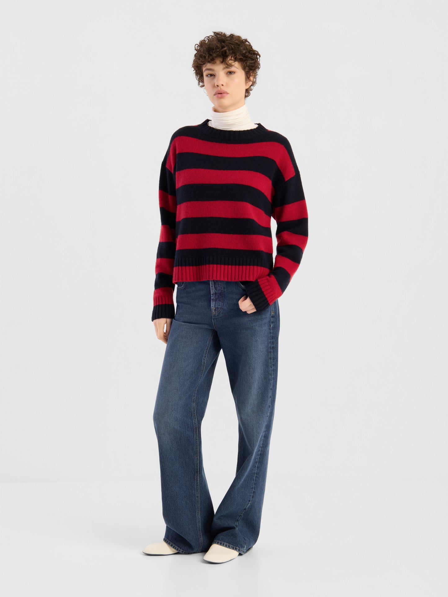 Eine Person, die den Pullover „Red Blue Stripes“ über einem weißen Rollkragenpullover trägt und dazu eine blaue Jeans mit weitem Bein sowie weiße Schuhe kombiniert hat, steht vor einem schlichten weißen Hintergrund.