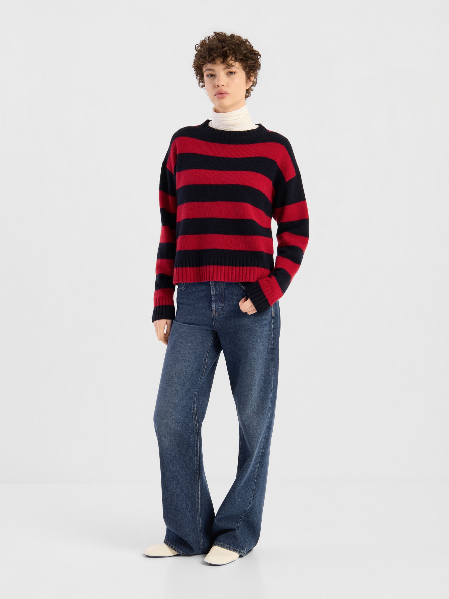 Eine Person, die den Pullover „Red Blue Stripes“ über einem weißen Rollkragenpullover trägt und dazu eine blaue Jeans mit weitem Bein sowie weiße Schuhe kombiniert hat, steht vor einem schlichten weißen Hintergrund.