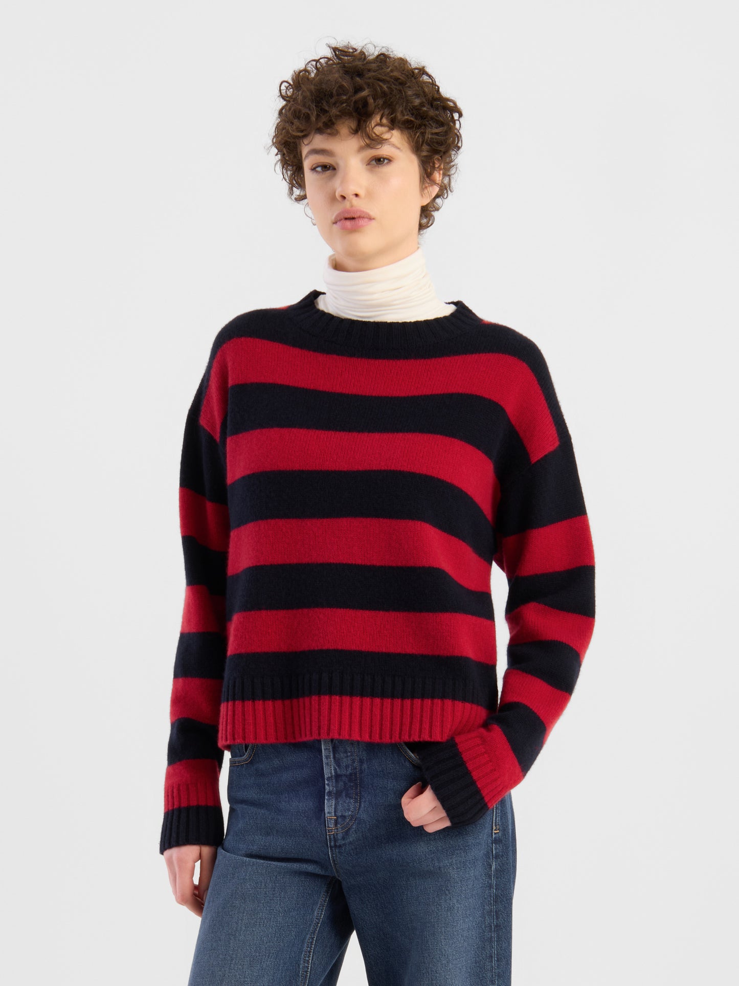 Eine Person, die den Pullover mit roten und blauen Streifen über einem weißen Rollkragenpullover und blauen Jeans trägt, steht vor einem schlichten, hellen Hintergrund.