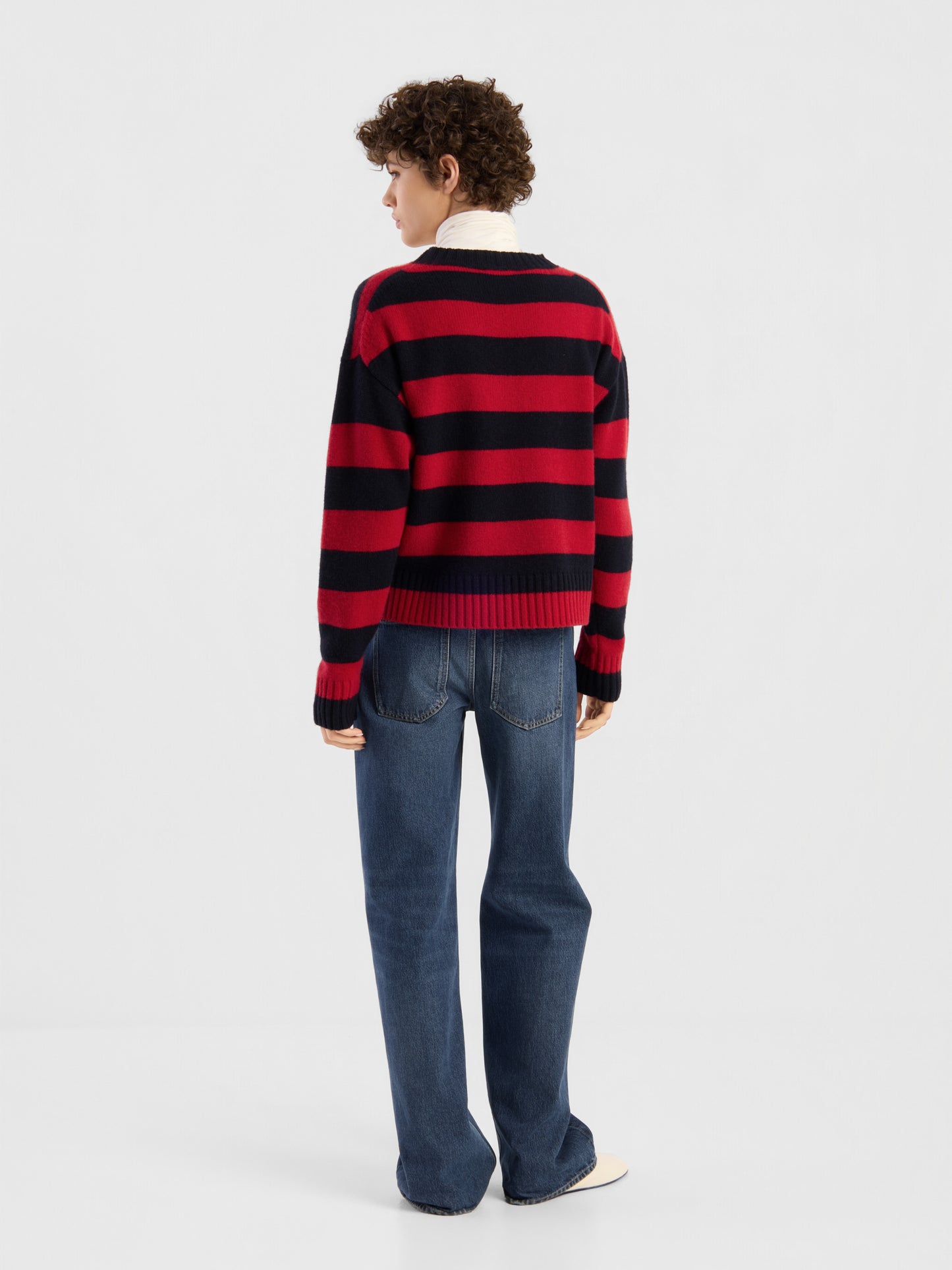 Eine Person, die mit dem Rücken zur Kamera steht und einen Pullover mit roten und blauen Streifen, eine blaue Jeans und weiße Schuhe trägt, vor einem schlichten weißen Hintergrund.