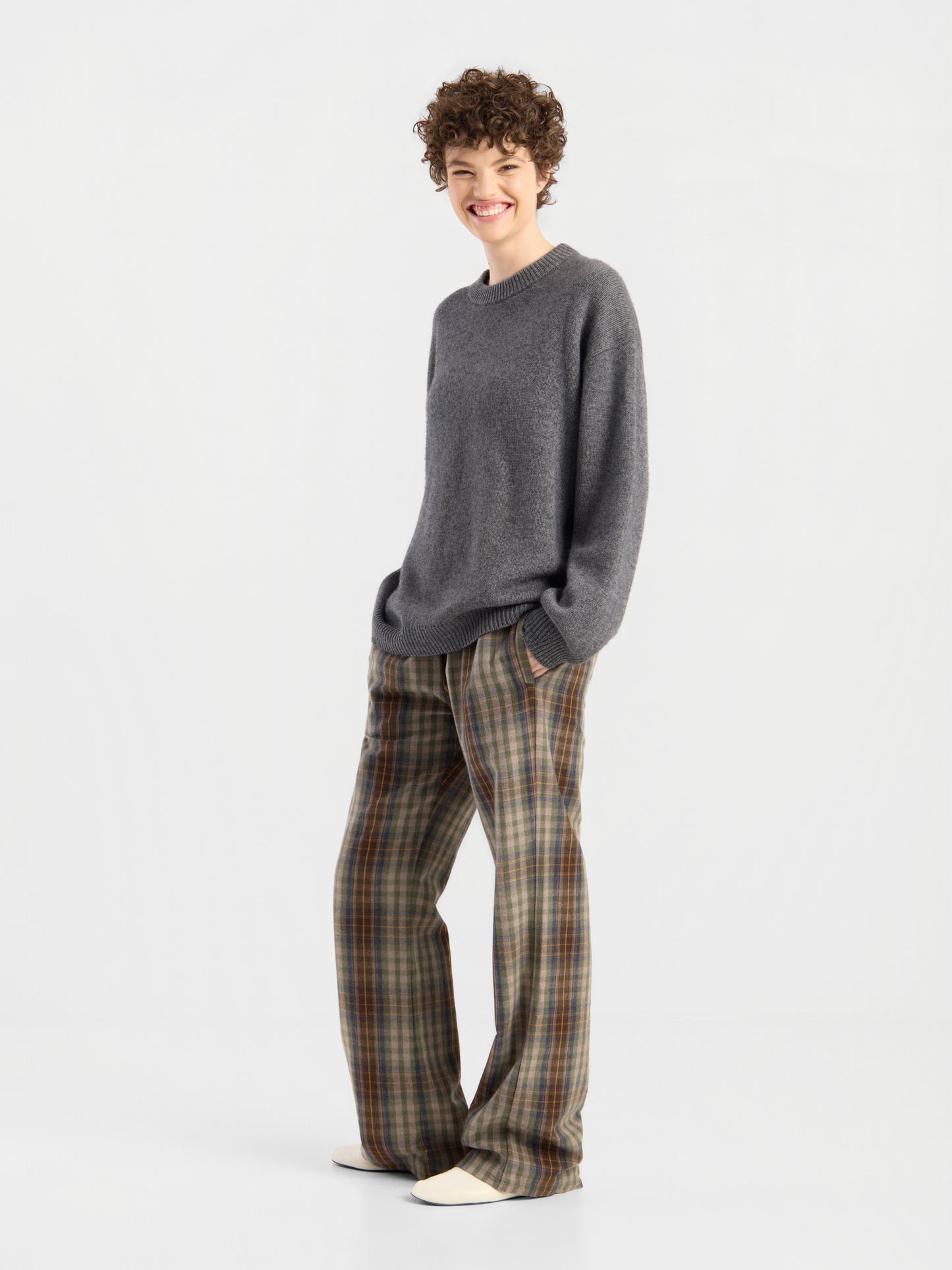 „Wearing the Pants Karoline Check“: Eine Person mit kurzen Locken und einem grauen Pullover lächelt in weißen Schuhen vor einem schlichten, hellen Hintergrund.
