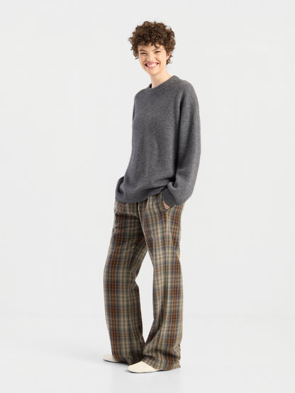 „Wearing the Pants Karoline Check“: Eine Person mit kurzen Locken und einem grauen Pullover lächelt in weißen Schuhen vor einem schlichten, hellen Hintergrund.