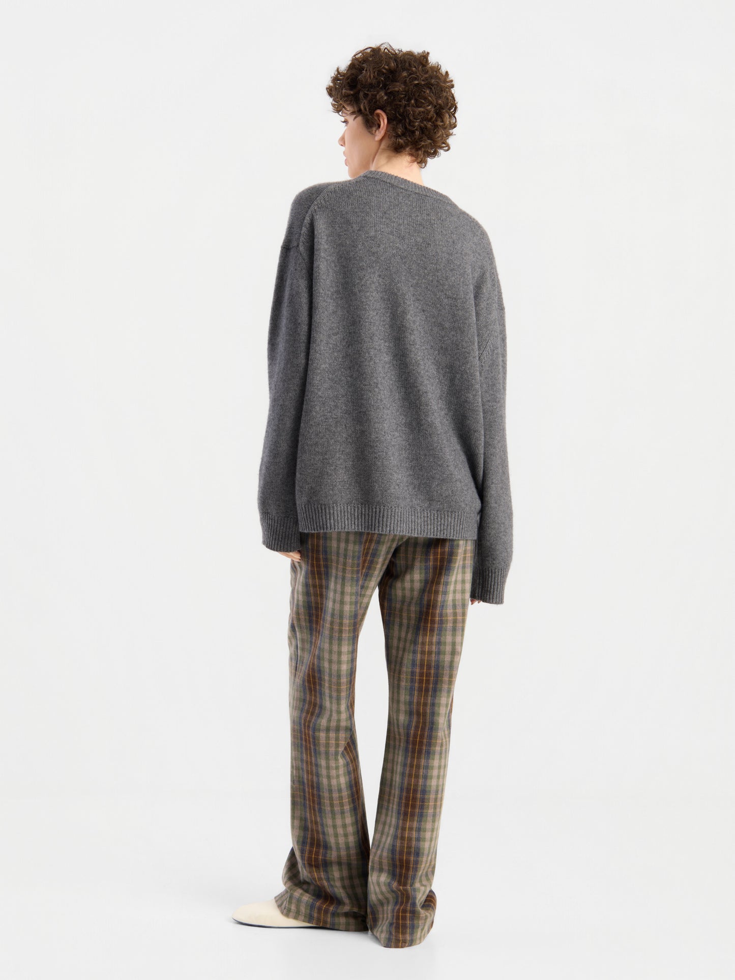 Eine Person mit lockigem Haar steht mit dem Rücken zur Kamera und trägt einen lockeren grauen Pullover sowie die Hose „Karoline Check“. Der schlichte weiße Hintergrund unterstreicht das lässige Outfit.