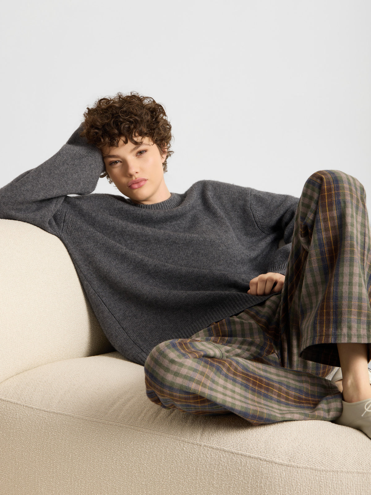 Eine Person mit kurzen Locken sitzt auf einem beigen Sofa. Sie trägt einen lockeren Pullover von „Pullover for Everybody Anthra“ und eine karierte Hose, hat einen Arm auf die Rückenlehne des Sofas gelegt und sieht entspannt aus.