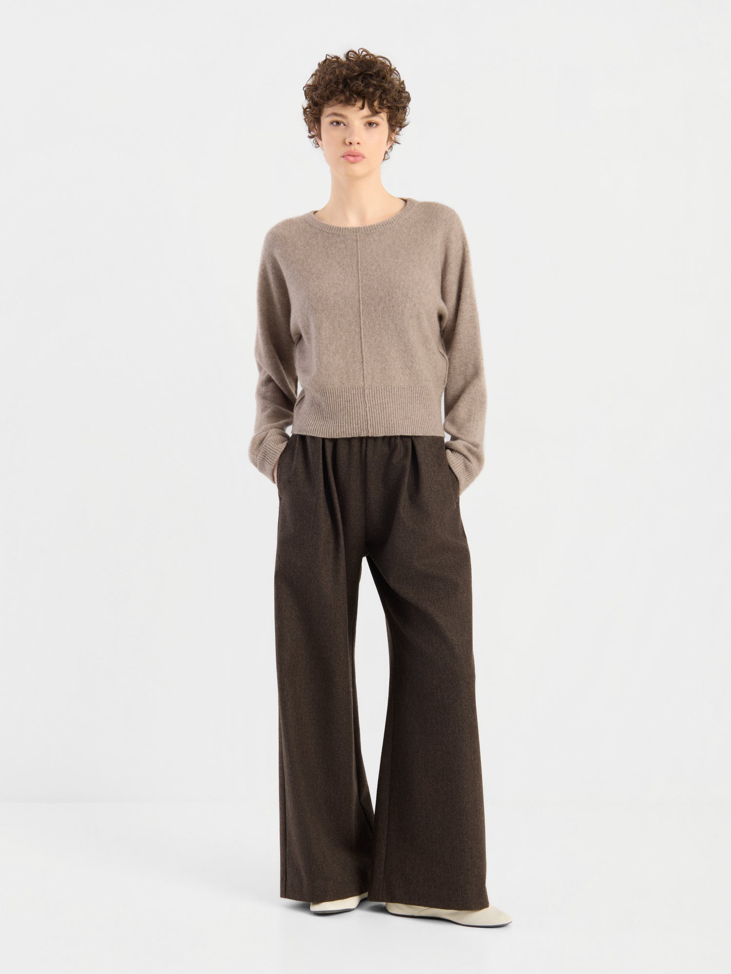 Eine Person mit kurzen Locken trägt den Pullover „Charly Beige“ und eine lockere, weit geschnittene braune Hose und steht mit den Händen in den Taschen vor einem schlichten weißen Hintergrund.