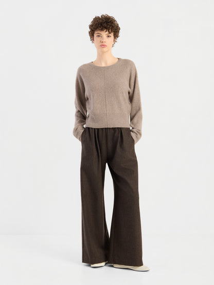 Eine Person mit kurzen Locken trägt den Pullover „Charly Beige“ und eine lockere, weit geschnittene braune Hose und steht mit den Händen in den Taschen vor einem schlichten weißen Hintergrund.