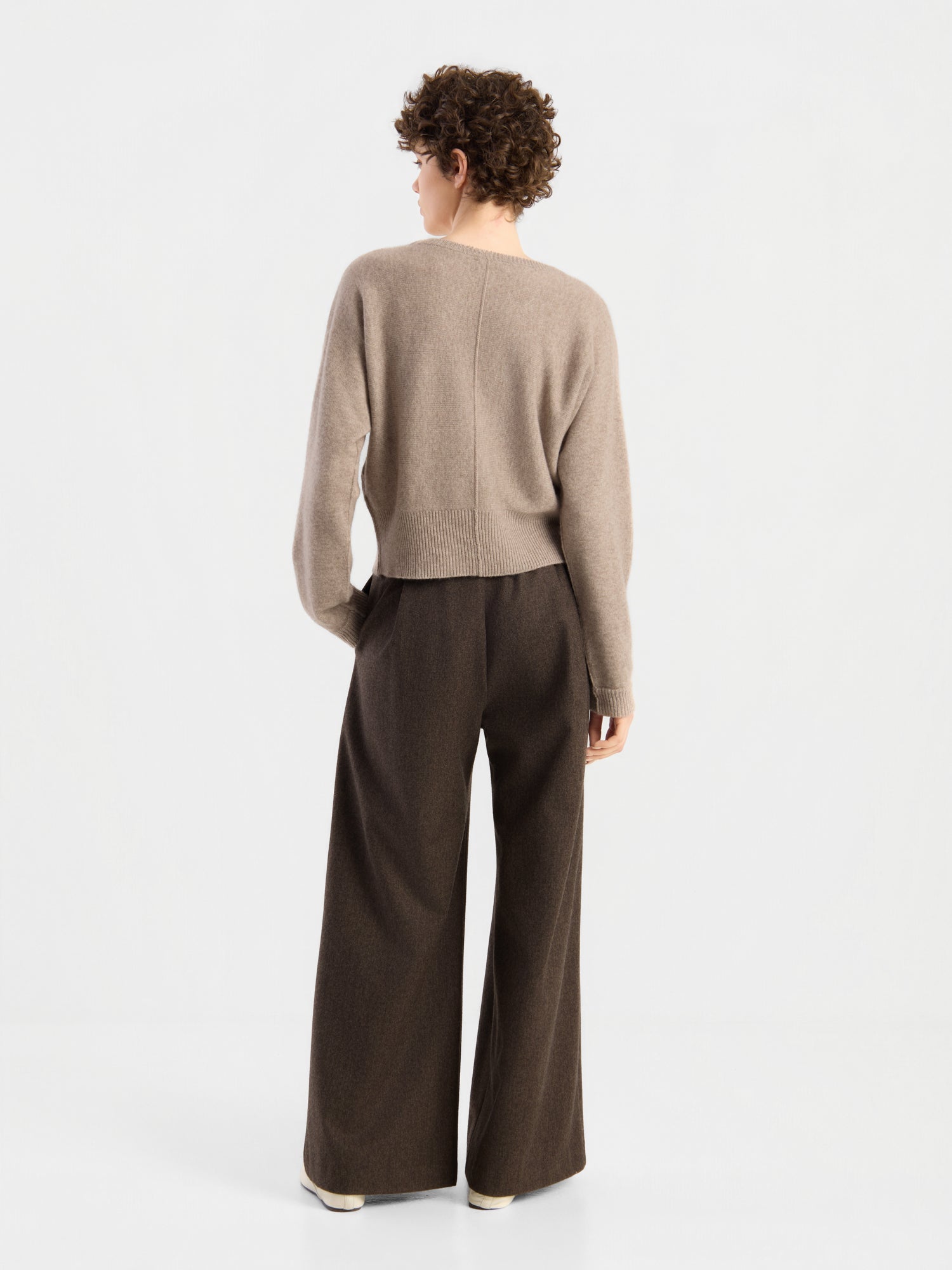 Eine Person mit kurzen Locken trägt den Pullover „Charly Beige“ und eine braune Hose mit weitem Bein und steht mit dem Rücken zur Kamera vor einem schlichten weißen Hintergrund.