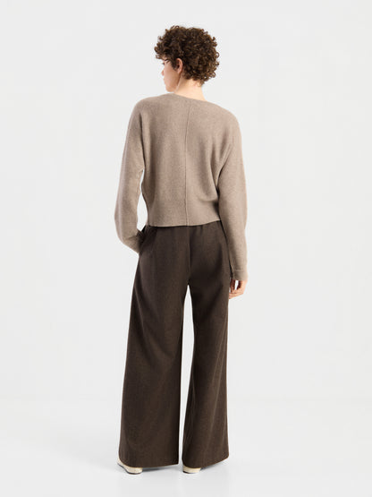Eine Person mit kurzen Locken trägt den Pullover „Charly Beige“ und eine braune Hose mit weitem Bein und steht mit dem Rücken zur Kamera vor einem schlichten weißen Hintergrund.