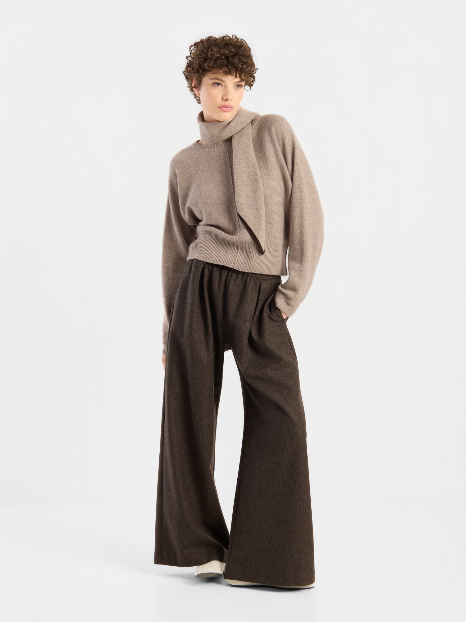 Eine Person mit kurzen Locken trägt den Pullover „Charly Beige“ mit asymmetrischem Kragen, dazu eine lockere, weit geschnittene braune Hose, und steht vor einem schlichten weißen Hintergrund.