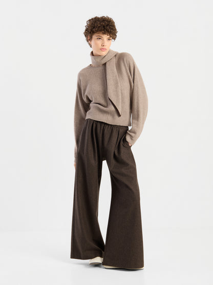 Eine Person mit kurzen Locken trägt den Pullover „Charly Beige“ mit asymmetrischem Kragen, dazu eine lockere, weit geschnittene braune Hose, und steht vor einem schlichten weißen Hintergrund.