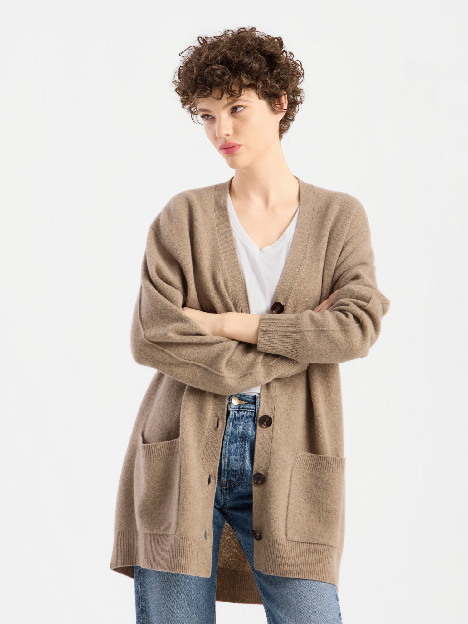 Eine Person mit kurzen Locken trägt einen beigen Cardigan „Lucia Beige“ über einem weißen Hemd und blauen Jeans und steht mit verschränkten Armen vor einem schlichten weißen Hintergrund.