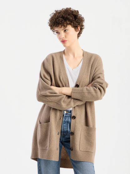 Eine Person mit kurzen Locken trägt einen beigen Cardigan „Lucia Beige“ über einem weißen Hemd und blauen Jeans und steht mit verschränkten Armen vor einem schlichten weißen Hintergrund.