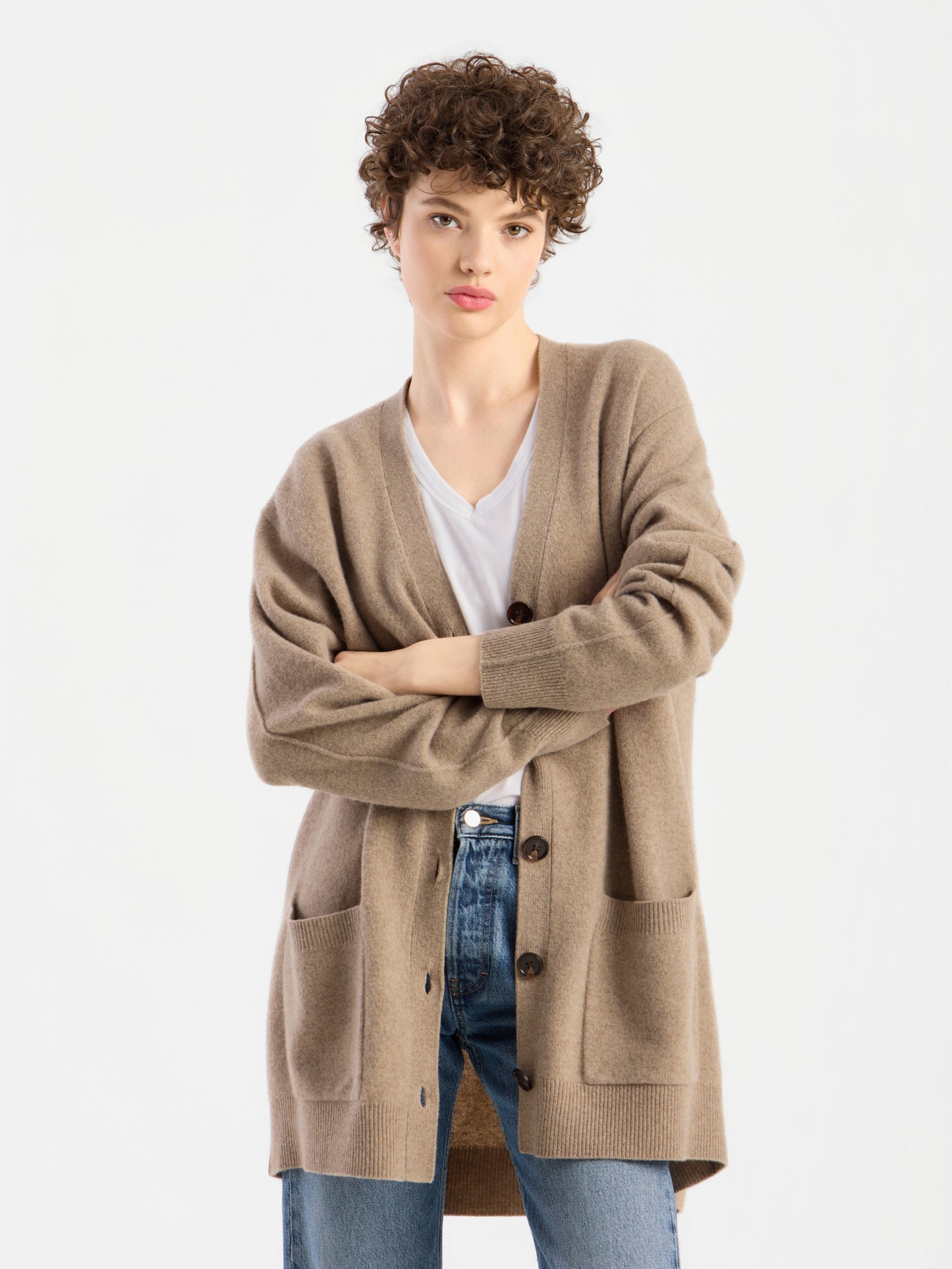 Eine Person mit kurzen Locken steht mit verschränkten Armen vor einem schlichten weißen Hintergrund und trägt den Cardigan „Lucia Beige“ über einem weißen Hemd und blauen Jeans.
