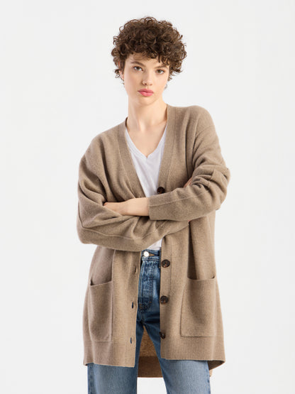 Eine Person mit kurzen Locken steht mit verschränkten Armen vor einem schlichten weißen Hintergrund und trägt den Cardigan „Lucia Beige“ über einem weißen Hemd und blauen Jeans.