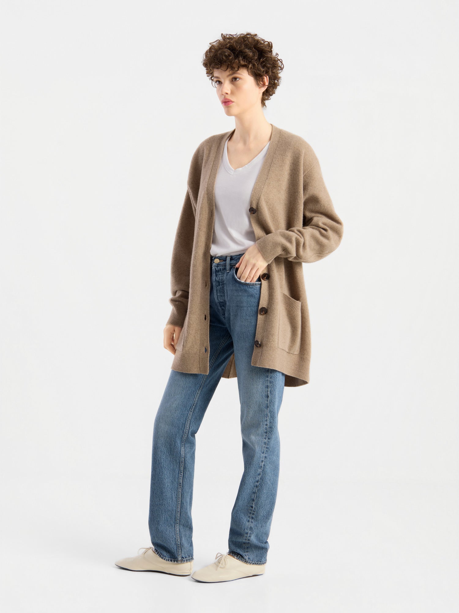 Eine Person mit kurzen Locken trägt einen hellbraunen Cardigan „Lucia Beige“ über einem weißen T-Shirt, blauen Jeans und beigen Schuhen und steht vor einem schlichten weißen Hintergrund.