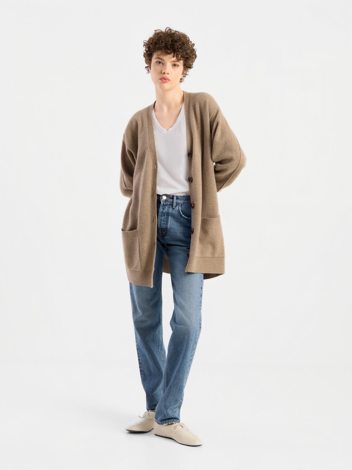 Eine Person mit kurzen Locken trägt den Cardigan Lucia Beige, einen Damen-Cardigan aus extrafeiner Merinowolle, über einer weißen Bluse, blauen Jeans und weißen Schuhen, während sie vor einem schlichten weißen Hintergrund steht.