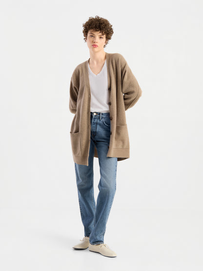 Eine Person mit kurzen Locken trägt den Cardigan Lucia Beige, einen Damen-Cardigan aus extrafeiner Merinowolle, über einer weißen Bluse, blauen Jeans und weißen Schuhen, während sie vor einem schlichten weißen Hintergrund steht.