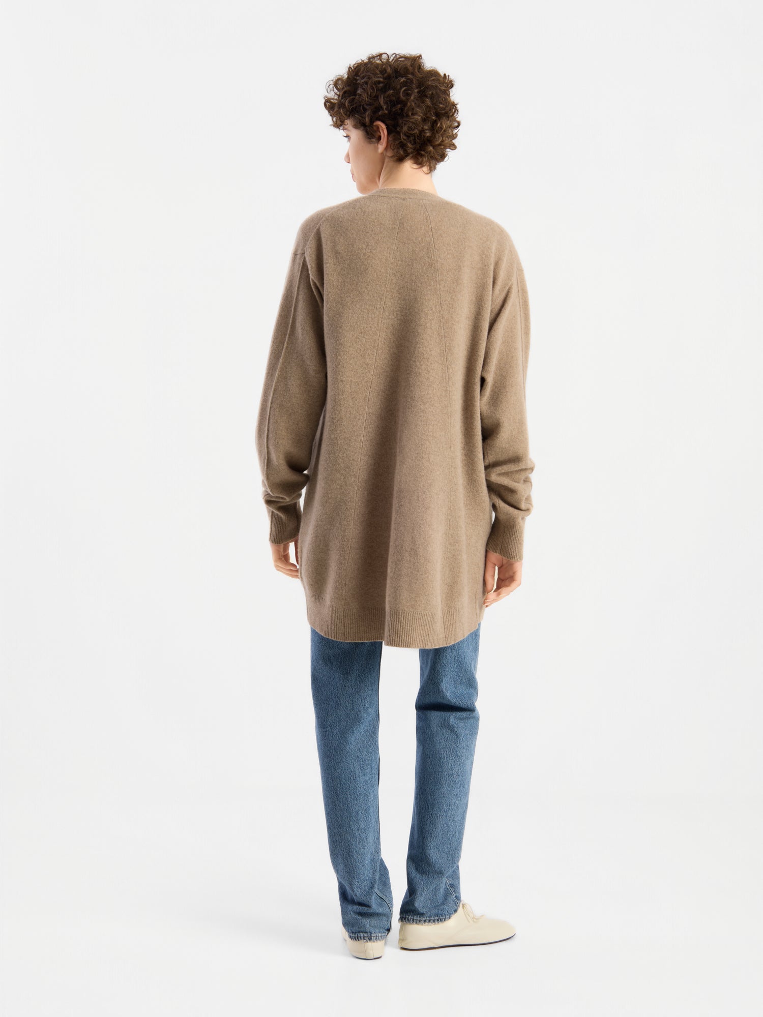 Eine Person mit kurzen Locken, bekleidet mit blauen Jeans und weißen Schuhen, die den langen beigen Cardigan „Lucia Beige“ trägt und vor einem schlichten weißen Hintergrund steht, mit dem Rücken zur Kamera.