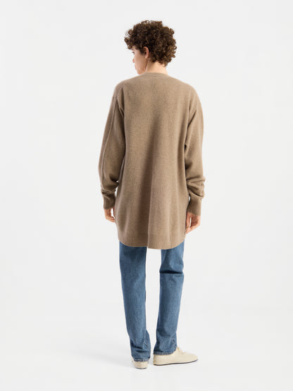 Eine Person mit kurzen Locken, bekleidet mit blauen Jeans und weißen Schuhen, die den langen beigen Cardigan „Lucia Beige“ trägt und vor einem schlichten weißen Hintergrund steht, mit dem Rücken zur Kamera.