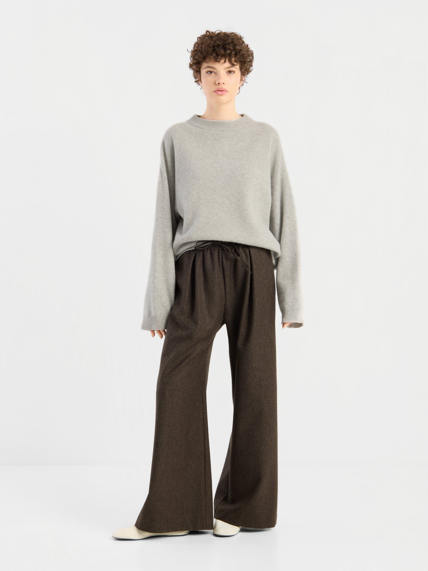 Eine Person mit kurzen Locken, die den Pullover „Liza“, einen lockeren grauen Pullover, eine braune Hose mit weitem Bein und weiße Schuhe trägt und vor einem schlichten weißen Hintergrund steht.