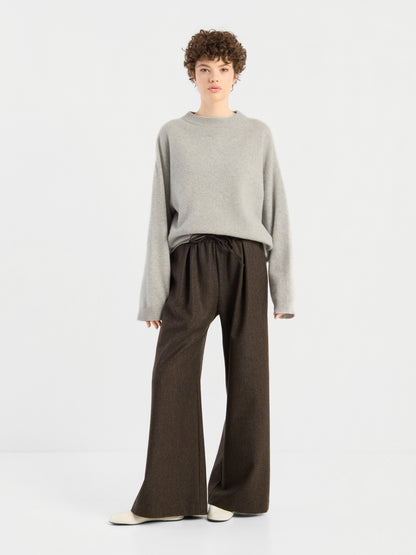 Eine Person mit kurzen Locken, die den Pullover „Liza“, einen lockeren grauen Pullover, eine braune Hose mit weitem Bein und weiße Schuhe trägt und vor einem schlichten weißen Hintergrund steht.
