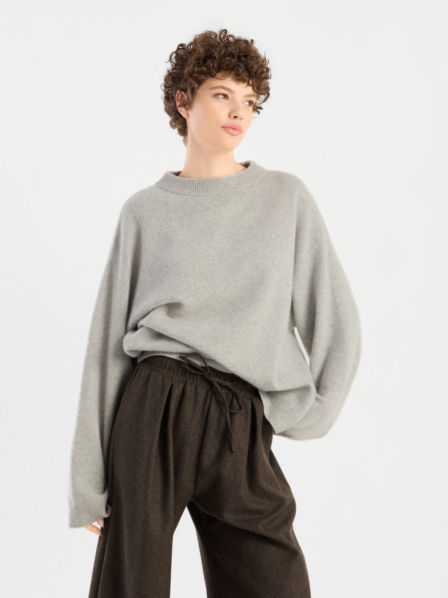 Eine Person mit kurzen Locken trägt den Pullover „Liza“, einen locker sitzenden hellgrauen Pullover, dazu eine dunkelbraune Hose mit hoher Taille. Sie steht vor einem schlichten weißen Hintergrund und blickt leicht zur Seite.