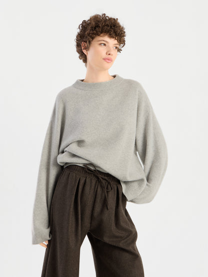 Eine Person mit kurzen Locken trägt den Pullover „Liza“, einen locker sitzenden hellgrauen Pullover, dazu eine dunkelbraune Hose mit hoher Taille. Sie steht vor einem schlichten weißen Hintergrund und blickt leicht zur Seite.