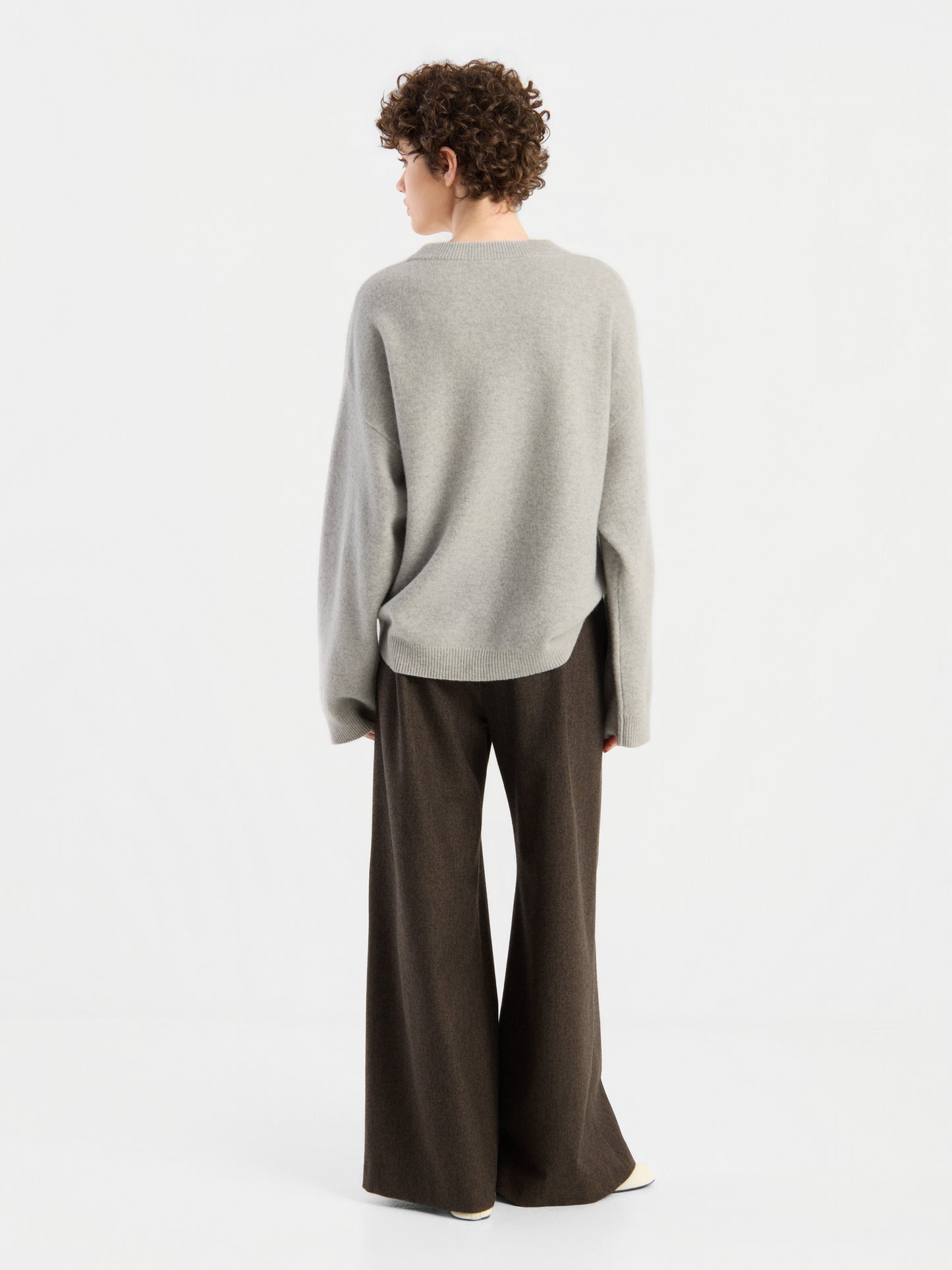 Eine Person mit kurzen Locken steht mit dem Rücken zur Kamera und trägt den Pullover „Liza“ – einen locker sitzenden grauen Pullover – sowie eine dunkelbraune Hose mit weitem Bein vor einem schlichten weißen Hintergrund.