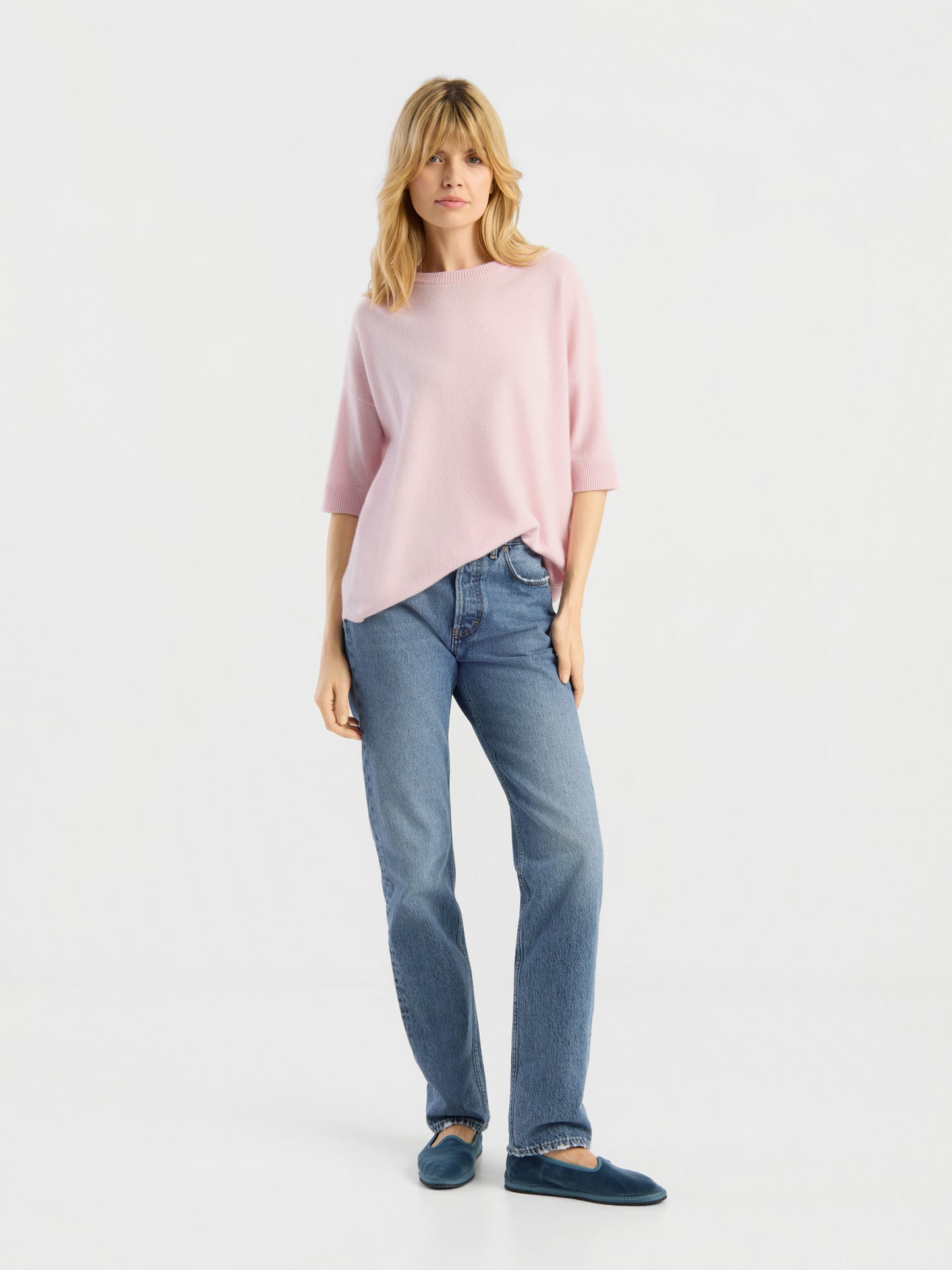 Vor einem schlichten weißen Hintergrund steht eine Frau mit blonden Haaren und einem entspannten Gesichtsausdruck. Sie trägt das Strick-T-Shirt „Aurora Rose“, eine blaue Jeans und dunkelblaue Hausschuhe.