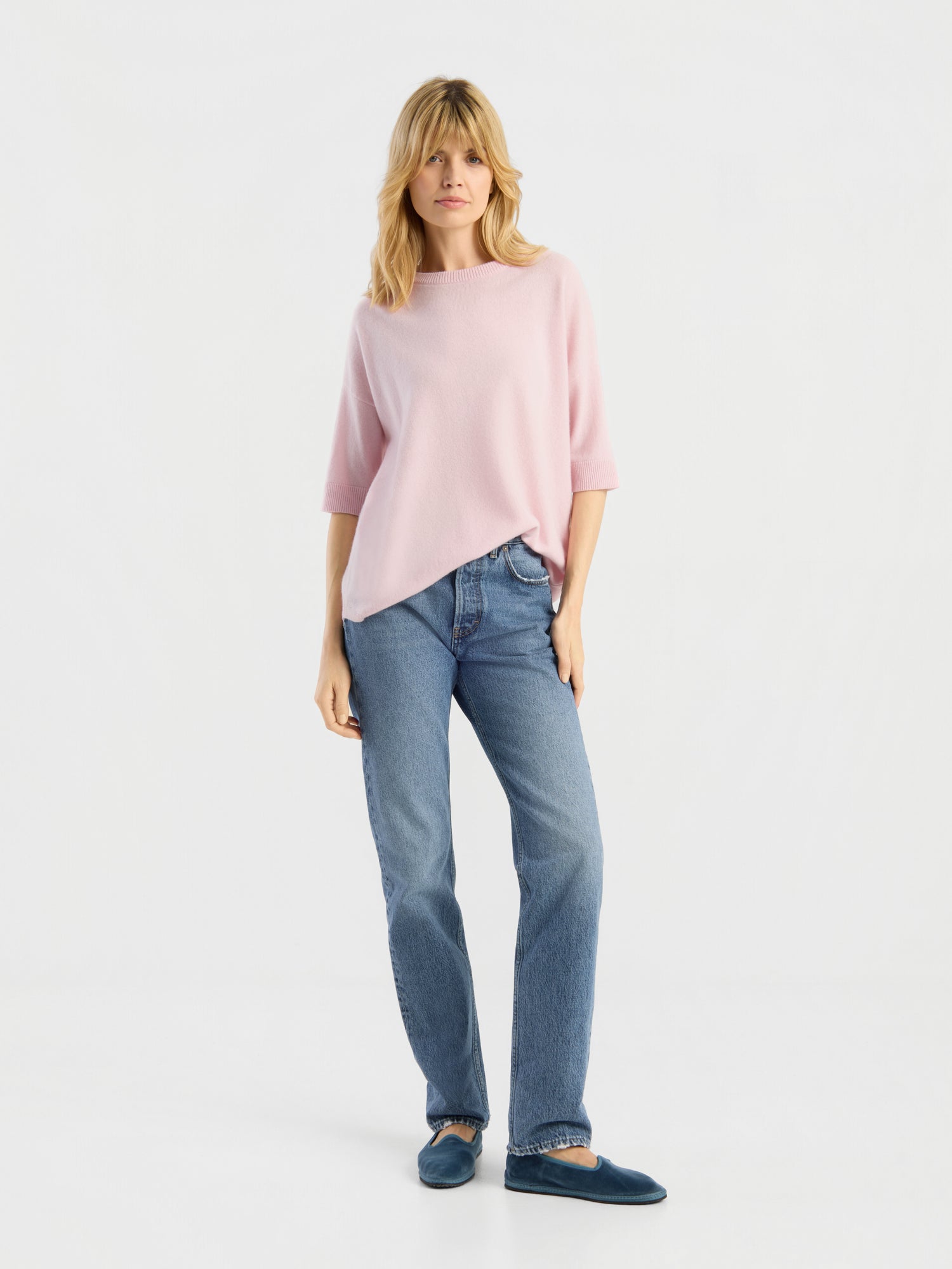 Vor einem schlichten weißen Hintergrund steht eine Frau mit blonden Haaren und einem entspannten Gesichtsausdruck. Sie trägt das Strick-T-Shirt „Aurora Rose“, eine blaue Jeans und dunkelblaue Hausschuhe.